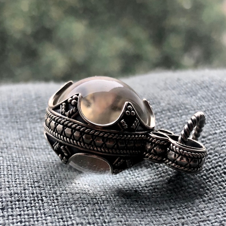 May include: A silver pendant featuring a clear quartz crystal sphere set within an intricate, ornate design. The pendant has a detailed, textured, silver chain.