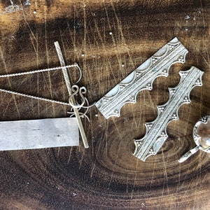 May include: A collection of silver jewellery making supplies on a wooden surface. The supplies include a silver chain, a silver bar, a silver piece with a decorative pattern, and a silver piece with a clear glass insert.