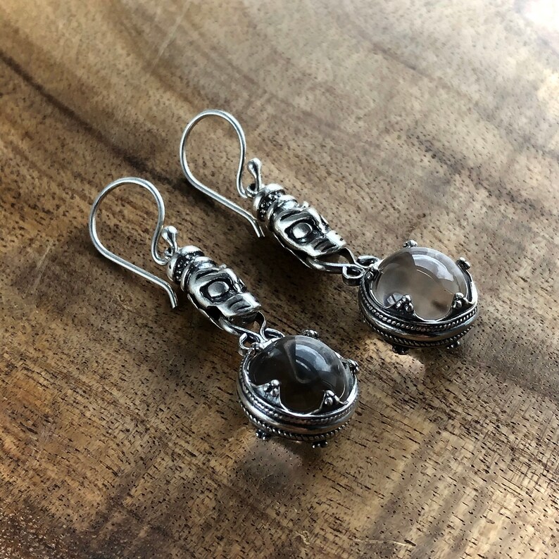 May include: A pair of silver dangle earrings with a clear and a smoky quartz stone. Each stone is set in a silver bezel with a decorative border. The earrings have a silver hook closure.