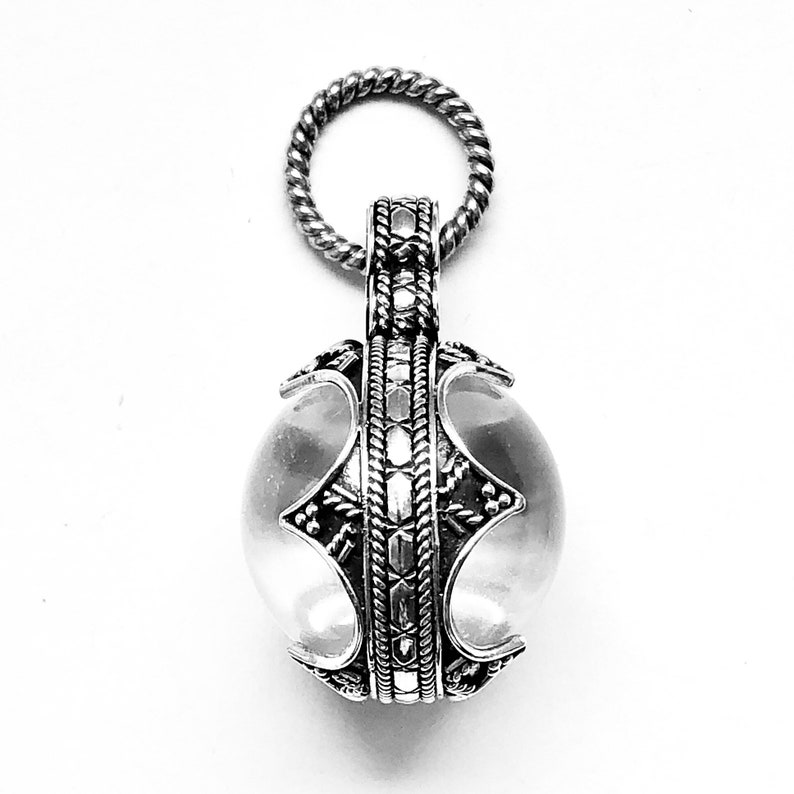 May include: A silver pendant featuring a clear crystal ball within. The pendant boasts a detailed design with intricate patterns and a twisted rope loop.