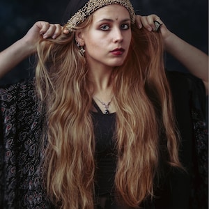 May include: A woman with long, wavy, light brown hair is wearing a black headscarf with gold beading and a black beaded necklace. She is wearing a black top with a floral pattern.