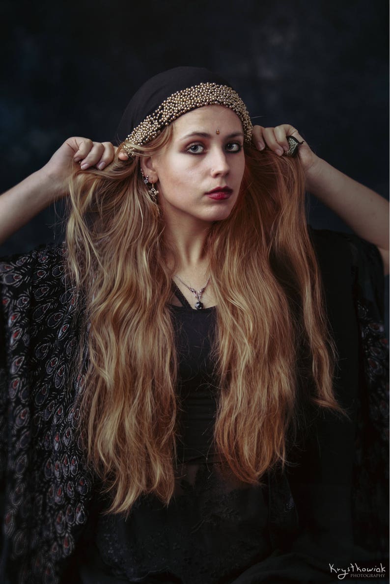 May include: A woman with long, wavy, light brown hair is wearing a black headscarf with gold beading and a black beaded necklace. She is wearing a black top with a floral pattern.