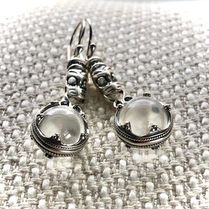 May include: Silver earrings with a clear stone and a detailed design. The earrings have a silver hook and a small silver charm.