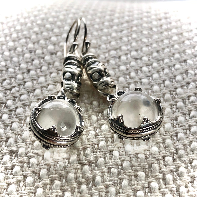 May include: Silver earrings with a clear stone and a detailed design. The earrings have a silver hook and a small silver charm.