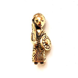 Könnte beinhalten: Ein goldfarbener Metallanhänger, der eine stilisierte Figur darstellt, die einen Schild und einen Speer hält. Die Figur hat ein einfaches, abgerundetes Gesicht mit geschlossenen Augen und einem kleinen Mund.