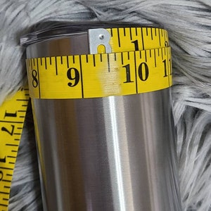 May include: A stainless steel tumbler with a black lid is measured with a yellow measuring tape. The tape shows a circumference of approximately 25 cm. The tumbler has a shiny, metallic finish.