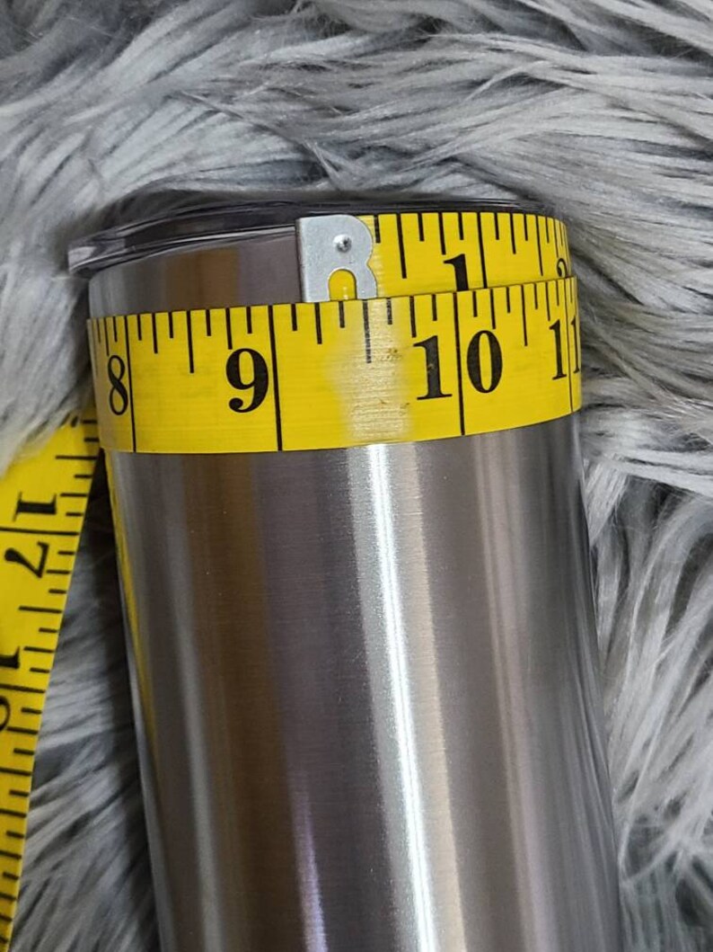 May include: A stainless steel tumbler with a black lid is measured with a yellow measuring tape. The tape shows a circumference of approximately 25 cm. The tumbler has a shiny, metallic finish.