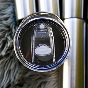 May include: Close-up of a stainless steel tumbler lid with a clear, sliding closure. The lid is black and has a clear plastic slider to open and close the drinking hole. Several other tumblers are visible in the background.