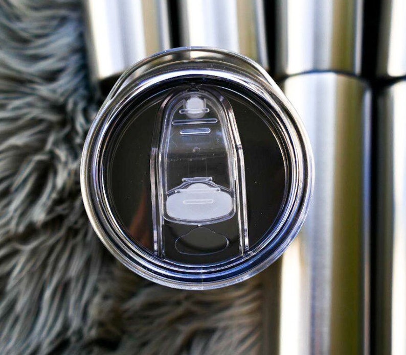 May include: Close-up of a stainless steel tumbler lid with a clear, sliding closure. The lid is black and has a clear plastic slider to open and close the drinking hole. Several other tumblers are visible in the background.