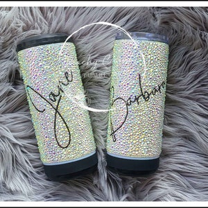 May include: Two stainless steel tumblers with black lids, covered in iridescent rhinestones and personalized with the names "Javi" and "Barbara" in black lettering.