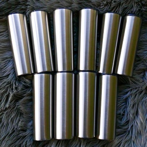 May include: Ten stainless steel tumblers with a brushed metal finish. Each tumbler has a cylindrical shape and a dark blue rim. The tumblers are arranged in two rows on a textured, grey surface.