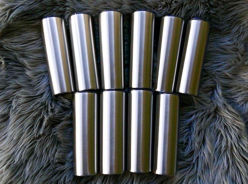 May include: Ten stainless steel tumblers with a brushed metal finish. Each tumbler has a cylindrical shape and a dark blue rim. The tumblers are arranged in two rows on a textured, grey surface.