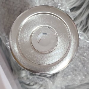 May include: A close-up of a shiny, silver metal lid. The lid features a circular design with concentric rings and a slightly raised centre. The surface has a brushed texture, reflecting light.