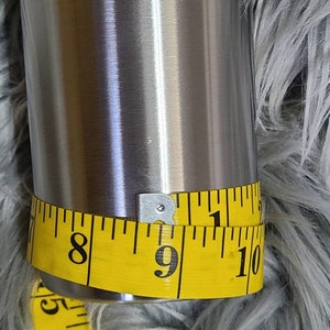 May include: A stainless steel tumbler with a yellow measuring tape wrapped around it. The tape indicates a circumference of approximately 23 cm. The tumbler has a shiny, metallic finish.