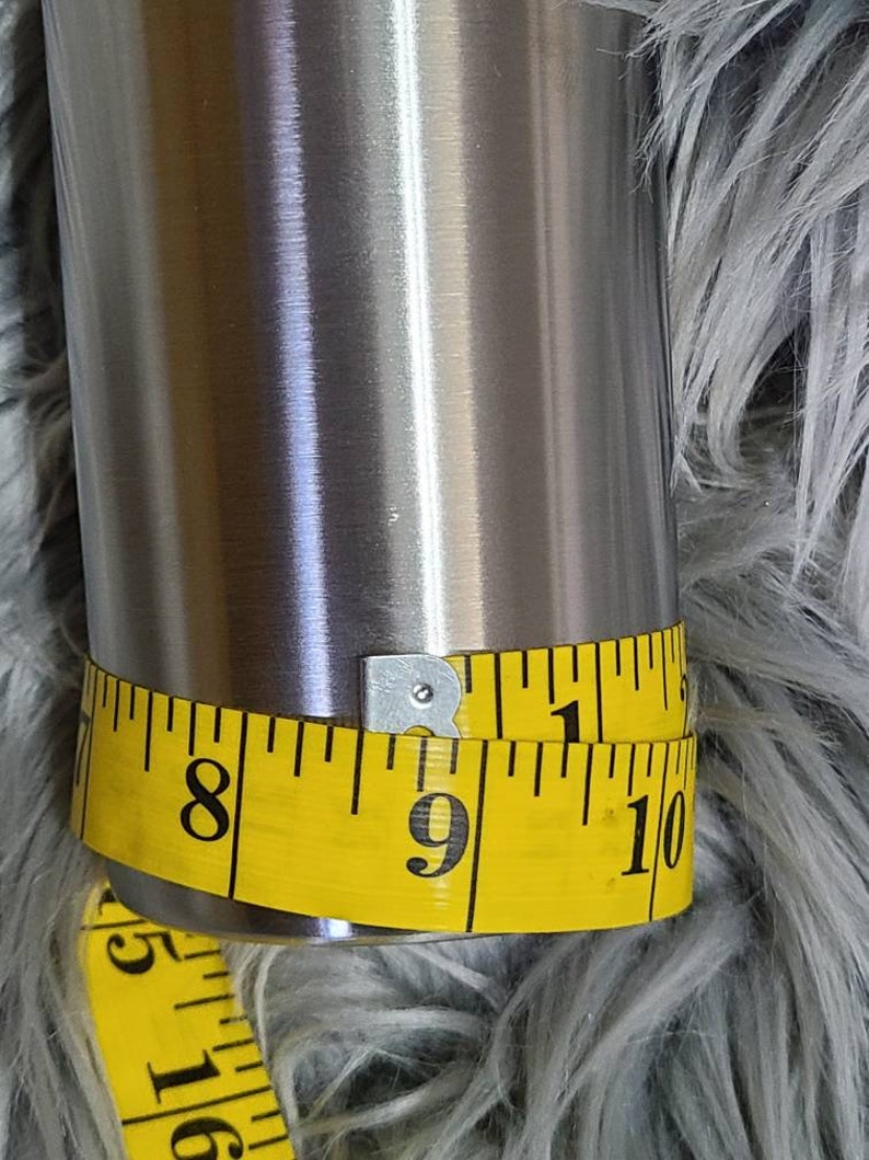 May include: A stainless steel tumbler with a yellow measuring tape wrapped around it. The tape indicates a circumference of approximately 23 cm. The tumbler has a shiny, metallic finish.