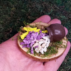 Peut inclure: Un plateau en bois miniature avec de la mousse et un petit pot brun débordant de perles jaunes, violettes et blanches.