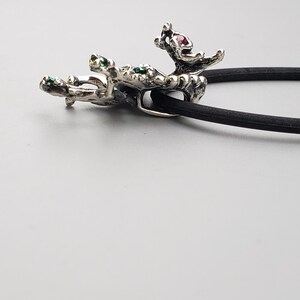 Op de afbeelding: Een zilverkleurige haarband met een decoratief, organisch gevormd accent. Het accent heeft groene en roze edelstenen. De haarband is zwart en ligt op een lichtgrijze ondergrond.