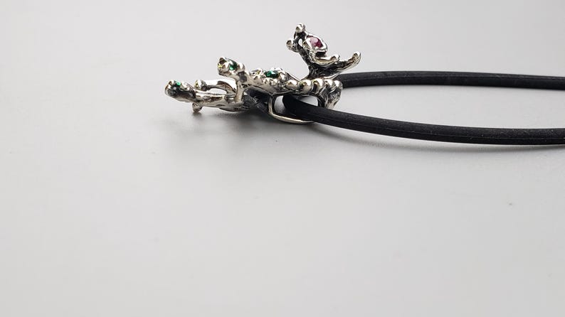Op de afbeelding: Een zilverkleurige haarband met een decoratief, organisch gevormd accent. Het accent heeft groene en roze edelstenen. De haarband is zwart en ligt op een lichtgrijze ondergrond.