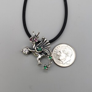 Op de afbeelding: Zilveren drakenhanger met rode, groene en heldere edelsteenaccenten. De draak hangt aan een zwart koord. Een Amerikaanse dime is naast de hanger ter referentie van de grootte.