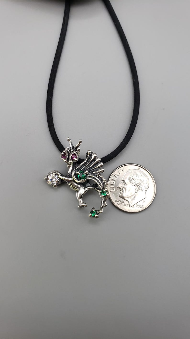 Op de afbeelding: Zilveren drakenhanger met rode, groene en heldere edelsteenaccenten. De draak hangt aan een zwart koord. Een Amerikaanse dime is naast de hanger ter referentie van de grootte.