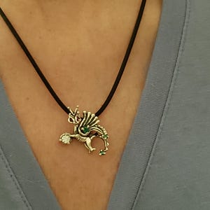 Op de afbeelding: Zilverkleurige drakenhanger aan een zwarte koordketting. De draak heeft groene en heldere edelsteenaccenten. De ketting wordt gedragen over een blauwe top.