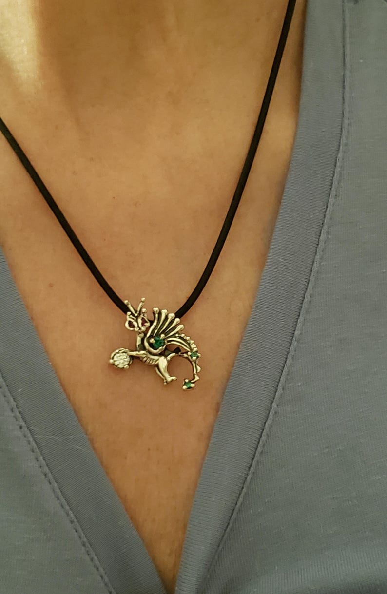 Op de afbeelding: Zilverkleurige drakenhanger aan een zwarte koordketting. De draak heeft groene en heldere edelsteenaccenten. De ketting wordt gedragen over een blauwe top.