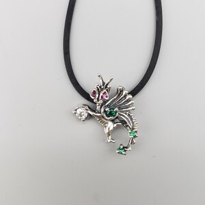 Op de afbeelding: Een zilveren drakenhangerketting met een zwart koord. De draak heeft rode, groene en heldere edelsteenaccenten. De hanger is gedetailleerd met vleugels en een gekrulde staart en hangt aan een zwart koord.