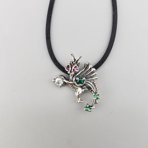 Op de afbeelding: Een zilveren drakenhangerketting met een zwart koord. De draak heeft rode, groene en heldere edelsteenaccenten. De hanger is gedetailleerd met vleugels en een gekrulde staart en hangt aan een zwart koord.