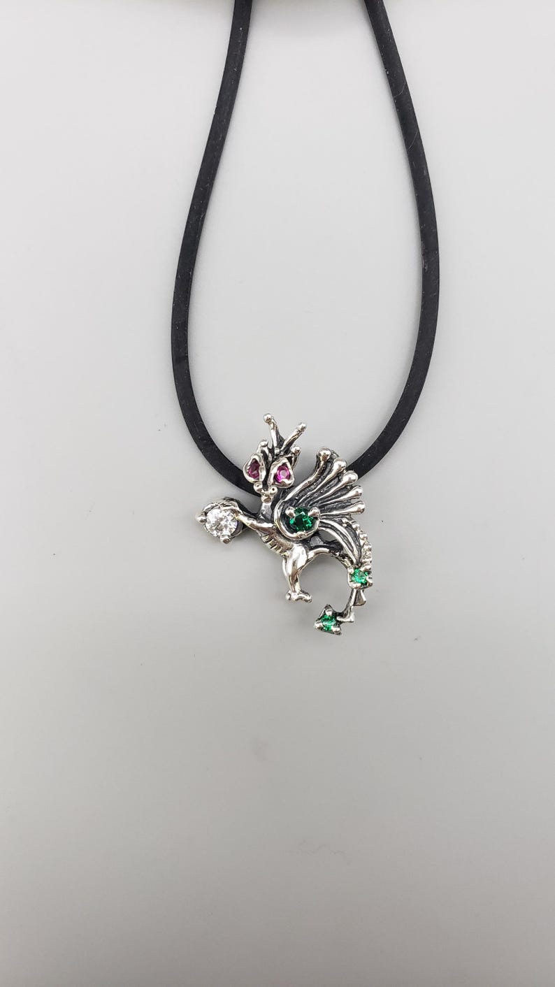 Op de afbeelding: Een zilveren drakenhangerketting met een zwart koord. De draak heeft rode, groene en heldere edelsteenaccenten. De hanger is gedetailleerd met vleugels en een gekrulde staart en hangt aan een zwart koord.