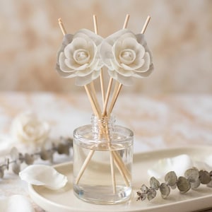 May include: A clear glass reed diffuser bottle containing a transparent liquid with several wooden reeds. Two large, white, multi-layered flower-shaped toppers are attached to the ends of the reeds, extending upward from the bottle opening.