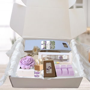 May include: A gift box filled with lavender-themed items. Includes a lavender-shaped soap, reed diffuser, wax melts, and a framed picture. The box is lined with white tissue paper, creating a visually appealing presentation.