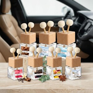 May include: A collection of seven square glass car air freshener bottles with light wood caps and two wooden diffuser sticks each. Each bottle features a unique label with text and imagery representing scents like cinnamon, fruit, and cotton.