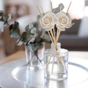 May include: A clear glass bottle with wooden sticks and white flower-shaped diffusers. The bottle is sitting on a silver tray with a sprig of eucalyptus in a glass vase.