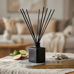 May include: A matte black square glass reed diffuser bottle with ten thin black reeds inserted. The diffuser is positioned on a circular tray featuring a split design of grey marble and brushed gold metal, accompanied by small citrus fruit slices.