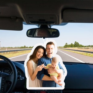 May include: A family of three, a man, a woman, and a baby, are smiling and looking at the camera. The man is holding the baby in his arms. The woman is standing next to the man and is holding the baby's hand. The family is in a car and the road is visible in the background.