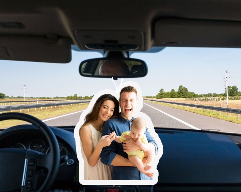 May include: A family of three, a man, a woman, and a baby, are smiling and looking at the camera. The man is holding the baby in his arms. The woman is standing next to the man and is holding the baby's hand. The family is in a car and the road is visible in the background.