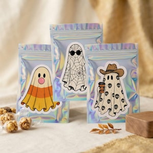 May include: Three iridescent holographic zipper pouches, each featuring a unique ghost sticker. One ghost resembles candy corn, one wears sunglasses over a spiderweb pattern, and one wears a cowboy hat while holding a pumpkin-themed coffee cup.