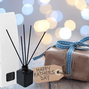 May include: A gift set for Father's Day, featuring a black diffuser with reeds, a wrapped present with a blue ribbon, and a gift tag that reads "Happy Fathers Day". A white box with reeds is also present.
