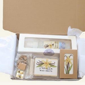 May include: Open cardboard box containing vanilla-scented products. Includes a room spray, wax melts, and decorative items. The wax melts are labeled "Vanilla Wax Melts" with a vanilla flower graphic. The box is lined with white tissue paper.