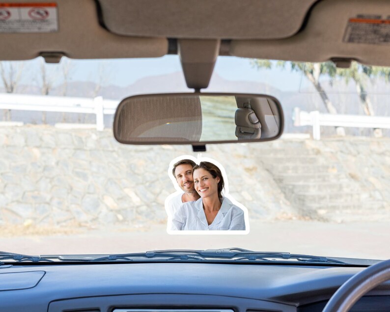 May include: A photo of a couple in a car. The couple is smiling and looking at the camera. The photo is attached to the rearview mirror of the car.