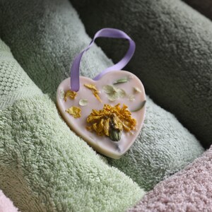 Puede incluir: Un jab&oacute;n rosa en forma de coraz&oacute;n con flores secas y una cinta lavanda. El jab&oacute;n est&aacute; sobre una pila de toallas.