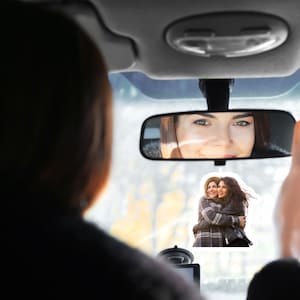 May include: A rearview mirror with a woman's reflection in it. A sticker of two women hugging is attached to the windshield.