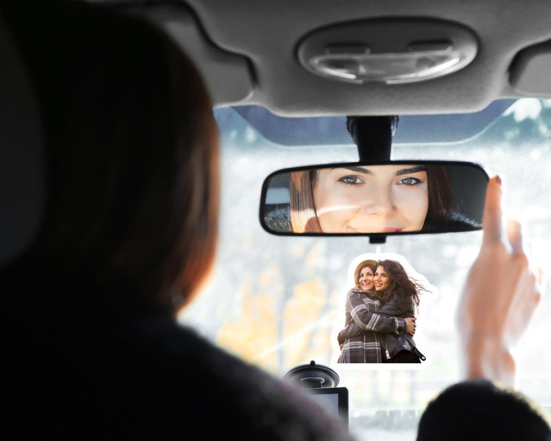 May include: A rearview mirror with a woman's reflection in it. A sticker of two women hugging is attached to the windshield.