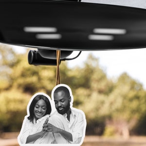 May include: Black and white photo of a couple in a car air freshener. The couple is smiling and looking at each other. The air freshener is hanging from the rearview mirror.