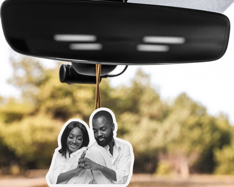 May include: Black and white photo of a couple in a car air freshener. The couple is smiling and looking at each other. The air freshener is hanging from the rearview mirror.