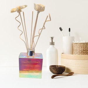 May include: A square iridescent glass diffuser bottle with a silver cap and wooden diffuser sticks topped with white ceramic flowers. The bottle is filled with a red and yellow liquid.