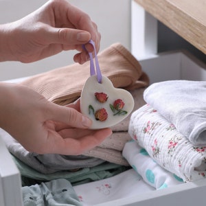 Puede incluir: Un derretido de cera blanco en forma de coraz&oacute;n con rosas rosadas secas y hojas verdes. El derretido de cera est&aacute; colgado de una cinta morada y est&aacute; siendo sostenido por una mano.