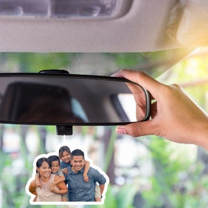 May include: A car air freshener with a photo of a family of four. The photo shows a man, a woman, and two children. The man is wearing a blue shirt and the woman is wearing a white shirt. The children are wearing colorful shirts. The air freshener is hanging from the rearview mirror of a car.