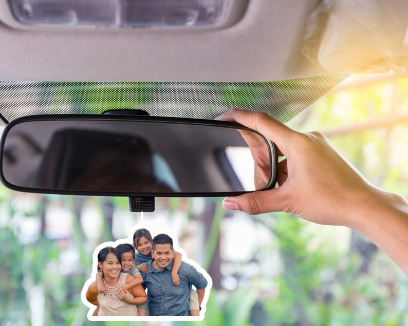 May include: A car air freshener with a photo of a family of four. The photo shows a man, a woman, and two children. The man is wearing a blue shirt and the woman is wearing a white shirt. The children are wearing colorful shirts. The air freshener is hanging from the rearview mirror of a car.