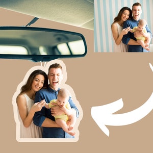 May include: A car air freshener with a photo of a family of three. The photo shows a man, a woman, and a baby. The man is holding the baby, and the woman is smiling at the camera. The air freshener is white and has a rounded shape.